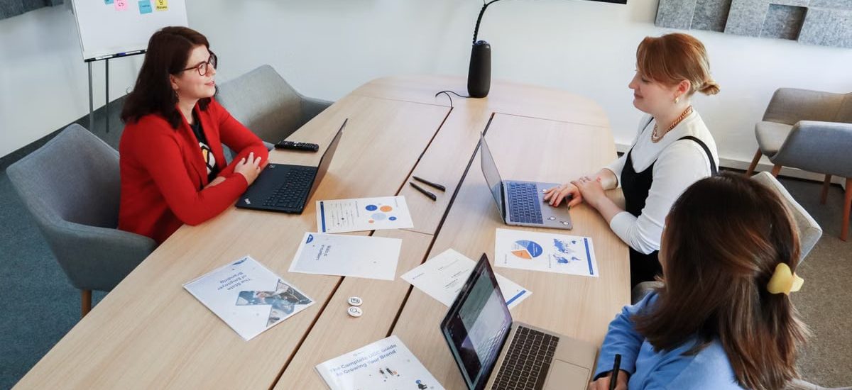 Three women in an office setting discussing business documents on laptops and paper.